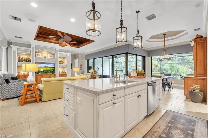 Kitchen island with light-toned cabinetry, quartz countertops, and stainless-steel dishwasher, complemented by three tiered metal pendant lights, wood-paneled tray ceiling detail, and large-format tile flooring