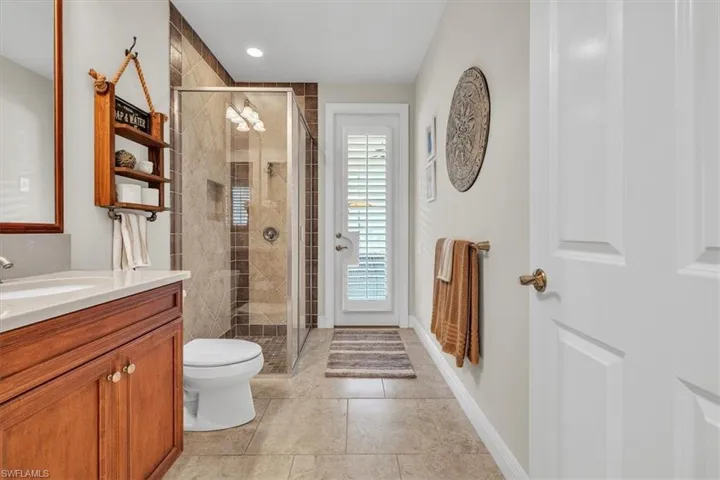 Bathroom featuring wood-finish cabinetry, neutral tile flooring, a glass-enclosed shower with ceramic tile surround, and an exterior door with plantation shutters