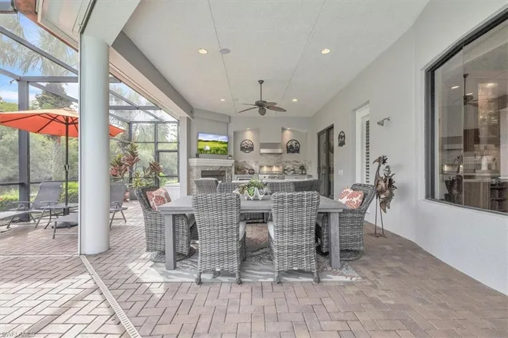 Covered outdoor patio featuring brick paver flooring, a stone-surround fireplace, a built-in outdoor kitchen with stainless steel appliances, a ceiling fan, and screened enclosure