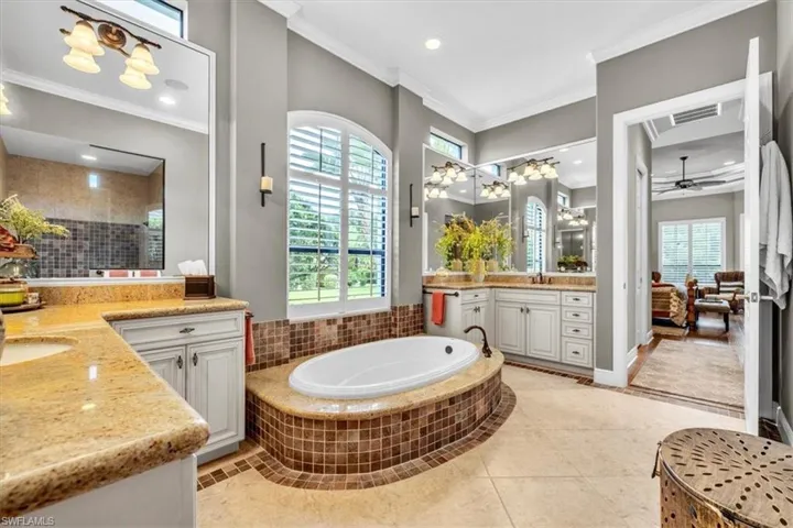 Bathroom featuring a soaking tub set in a tile-surround pedestal, light-toned cabinetry, natural stone countertops, arched window, and large wall mirrors