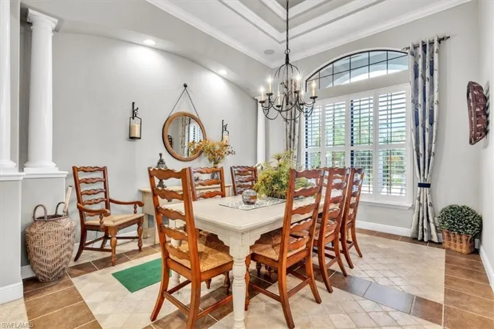 Dining room featuring a coffered ceiling, iron chandelier, arched window with plantation shutters, tiled flooring with a decorative border, and classic architectural columns