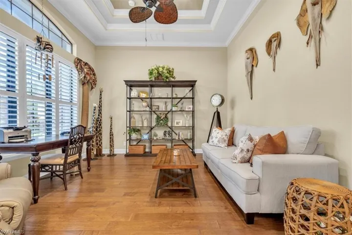 Tray ceiling with a central ceiling fan, expansive windows with plantation shutters, wood-finish flooring, and built-in metal shelving unit
