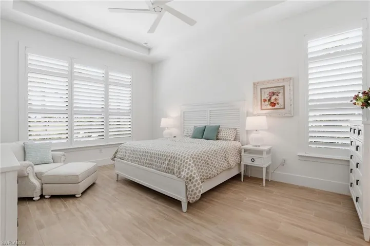 Spacious bedroom featuring wood-finish flooring, white plantation shutters, a tray ceiling, and a ceiling fan