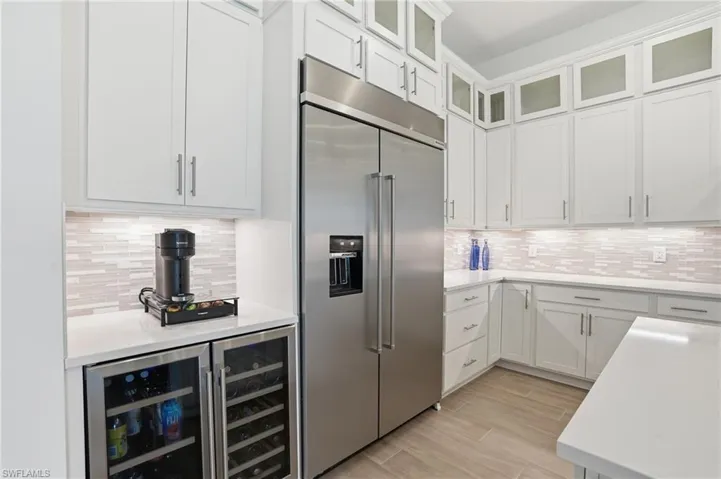 Gourmet kitchen with stainless steel refrigerator, white cabinetry, integrated beverage and wine coolers, light-toned tile backsplash, and light-colored countertops