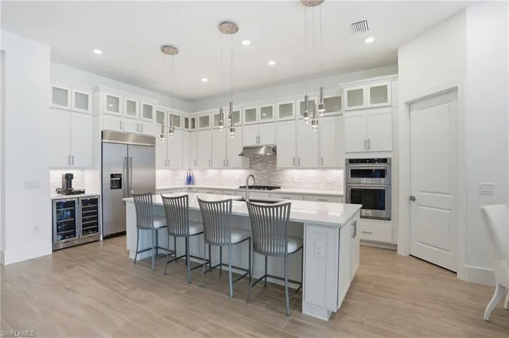 Spacious kitchen featuring an expansive island with seating, stainless steel appliances, white cabinetry with glass-front uppers, a tiled backsplash, and wood-finish flooring