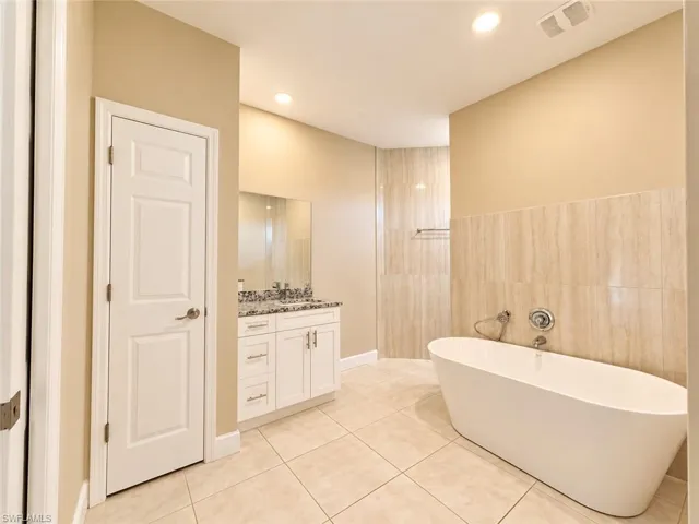 Freestanding soaking tub, tiled accent wall, dual vanity with undermount sinks, and tile flooring
