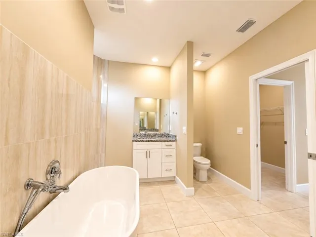 Freestanding soaking tub with chrome fixtures, tiled shower surround, dual-sink vanity with stone countertop, private commode, and tile flooring