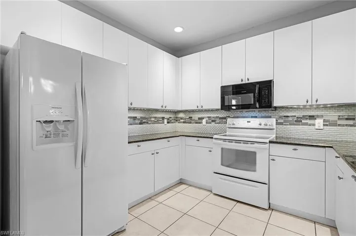 Kitchen featuring white cabinetry, dark countertops, a mosaic tile backsplash, white appliances, and light-colored tile flooring