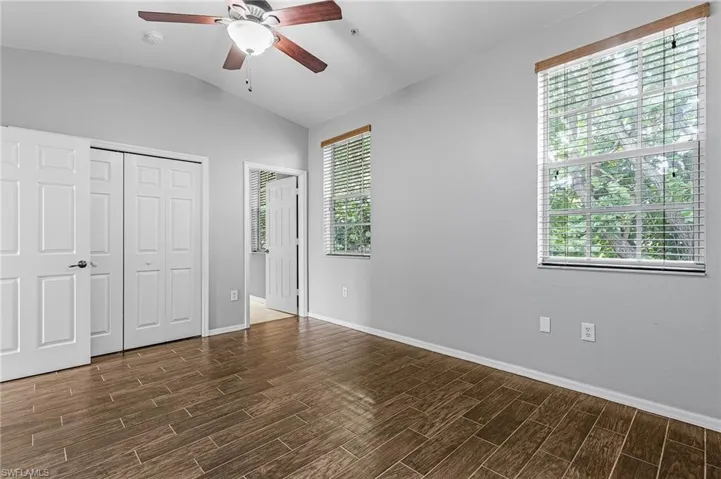 Interior room featuring wood-finish flooring, two windows with blinds, light gray walls, a ceiling fan with integrated lighting, and two white bi-fold closet doors