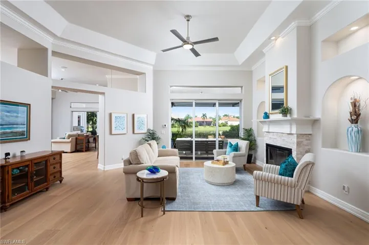 Living room featuring light wood finished floors, a tray ceiling, a stone fireplace, ceiling fan, and crown molding