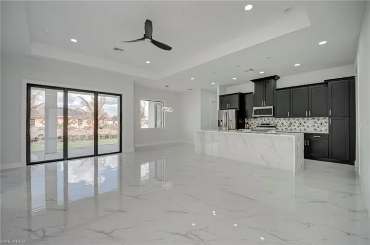Kitchen with a tray ceiling, dark cabinetry, a ceiling fan, a center island with sink, and light stone countertops