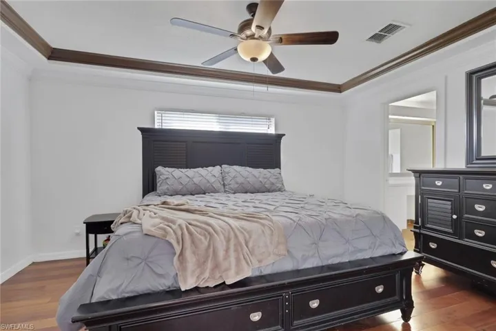 Bedroom with ornamental molding, a raised ceiling, dark wood finished floors, ensuite bath, and ceiling fan