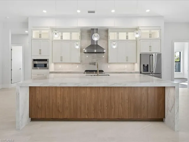 Kitchen with glass insert cabinets, light stone counters, a large island with sink, wall chimney exhaust hood, and stainless steel appliances
