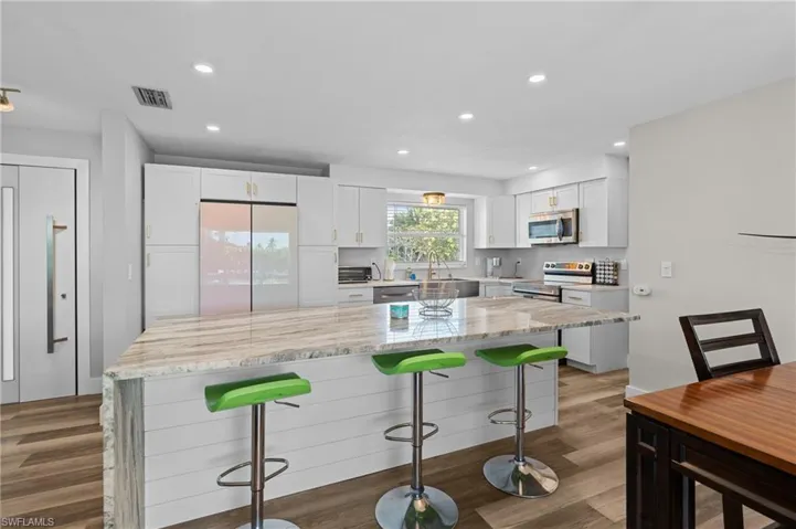 Kitchen with white cabinets, stainless steel appliances, visible vents, light stone countertops, and a breakfast bar area  - Virtually Edited Image