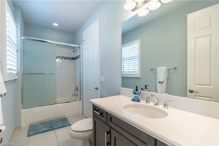 Bathroom featuring a vanity with an undermount sink and light-toned countertop, a shower-tub combination with sliding frosted glass doors, white subway tile surround, and a window with plantation shutters