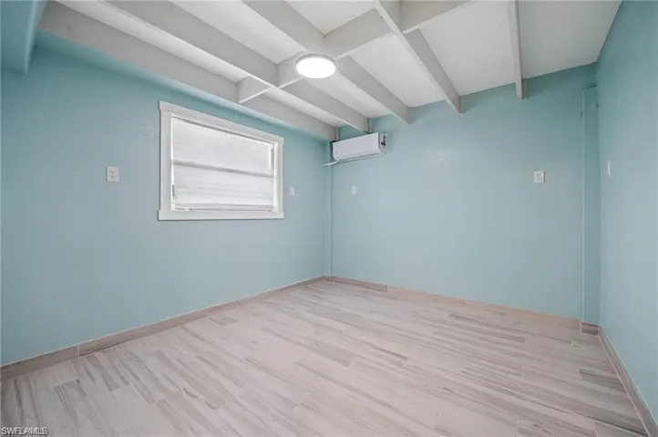 Empty room with a wall mounted AC, beamed ceiling, and light wood-type flooring