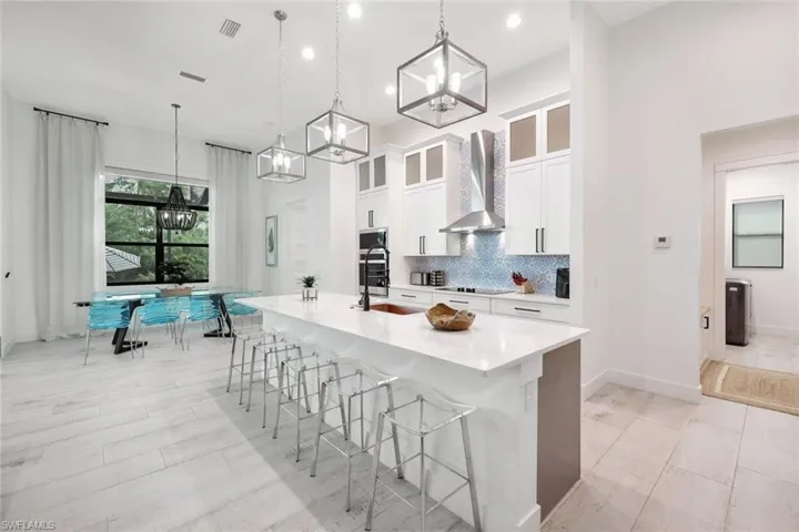 Kitchen featuring an island with sink, white cabinets, wall chimney range hood, appliances with stainless steel finishes, and decorative light fixtures