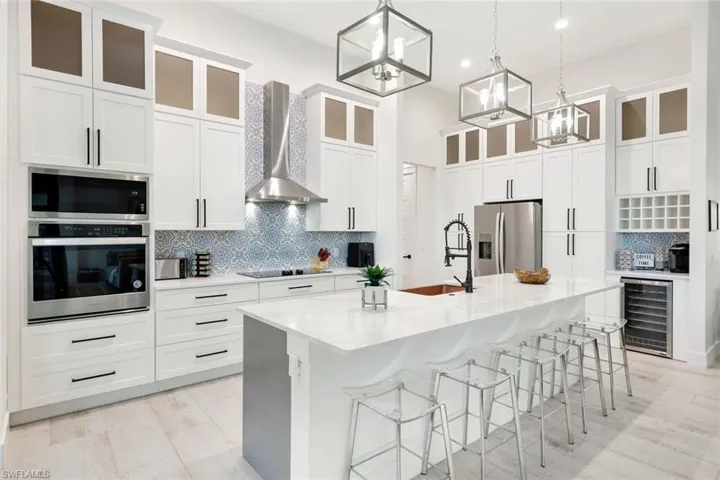 Kitchen featuring beverage cooler, wall chimney range hood, an inviting chandelier, stainless steel appliances, and a center island with sink