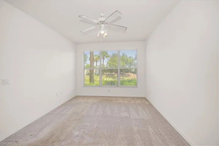 featuring neutral carpet flooring, a large window with horizontal blinds overlooking exterior trees, and a ceiling fan with integrated lighting