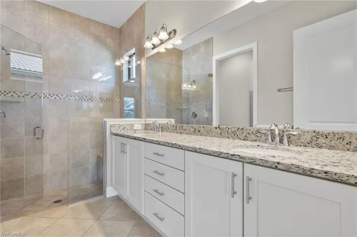 Primary bathroom with a large tiled walk-in shower with listello and frameless glass door, double vanity with granite counters and light tile floors.
