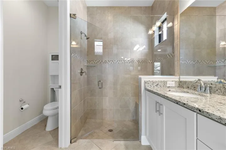 Primary bathroom with a private water closet, large tiled walk-in shower with listello and frameless glass door, double vanity with granite counters and light tile floors.