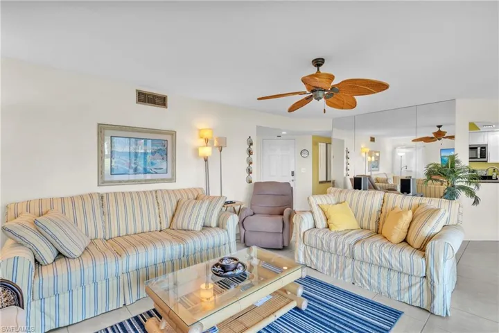 Bright and airy living room showcasing dual striped upholstered seating arrangements, ceiling fan with wood-finish blades, large windows with natural light, wood-finish coffee table, and an open floor plan connecting to the kitchen and dining areas