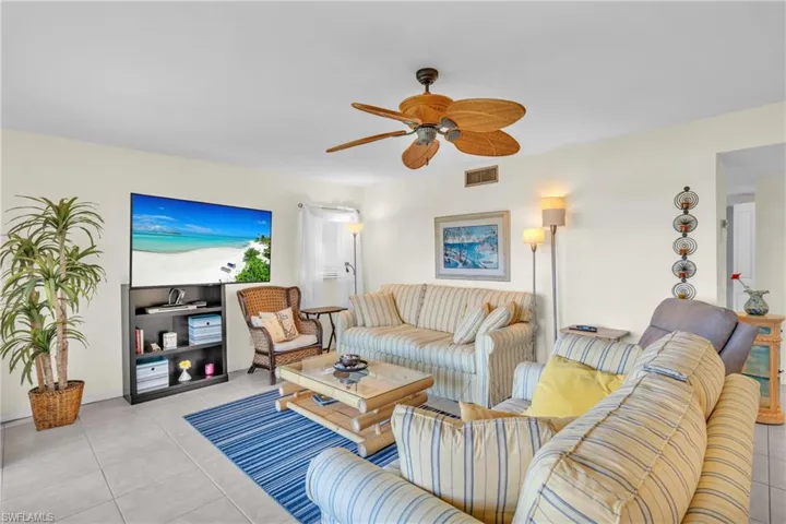 Bright and open living area featuring striped upholstered seating, wood-finish ceiling fan with light fixture, tile flooring, built-in media console with shelving, and large flat-screen television