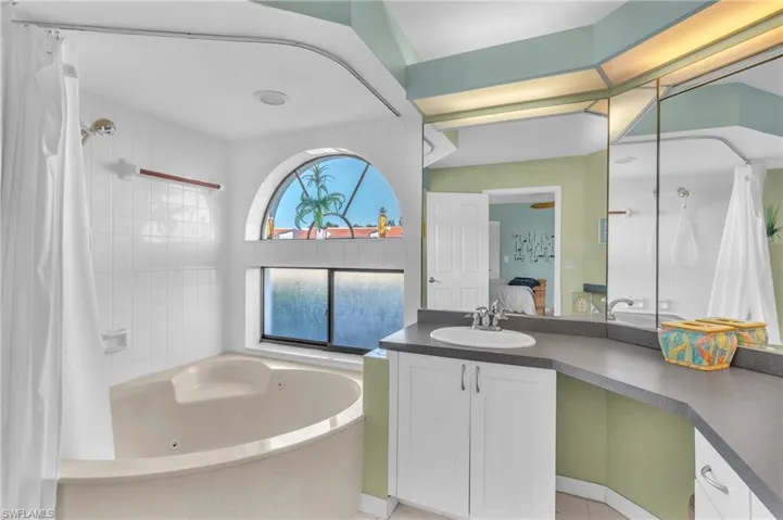 Spa-inspired primary bathroom featuring a curved soaking tub with integrated jets, dual vanity with gray quartz countertops and white cabinetry, frameless glass shower enclosure with subway tile surround, arched transom window with coastal views, and soft sage-green accent walls with recessed lighting