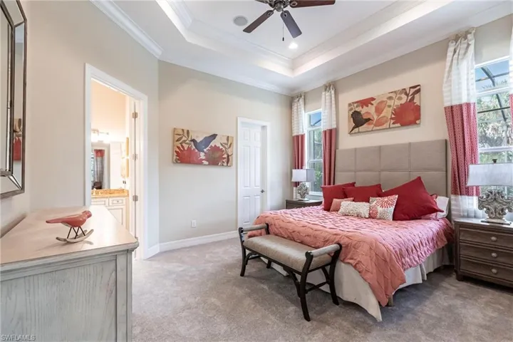 Bedroom with light colored carpet, baseboards, a tray ceiling, ensuite bath, and crown molding, walk in closet