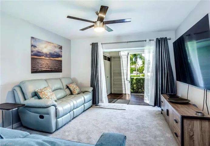 Living area featuring light-toned carpeting, a ceiling fan with integrated lighting, and a wall-mounted flat-screen display