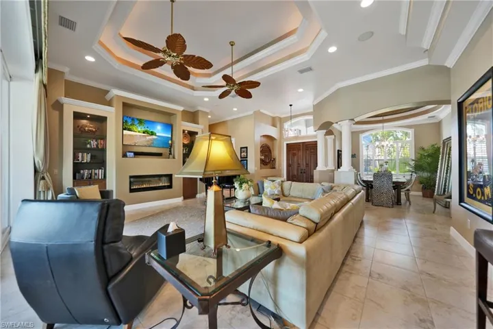 Tiled living room with ceiling fan with notable chandelier, decorative columns, a tray ceiling, and crown molding