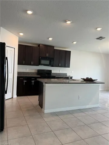 Kitchen featuring a textured ceiling, black appliances, an island with sink, light tile patterned floors, and dark stone countertops