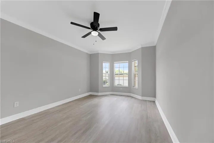 Spacious room featuring a bay window with three sashed windows, light gray walls, wood-finish flooring, white crown molding, and a black ceiling fan