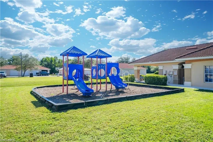 Outdoor recreational area featuring a blue and yellow playground structure with slides and climbing elements, set on a wood chip surface
