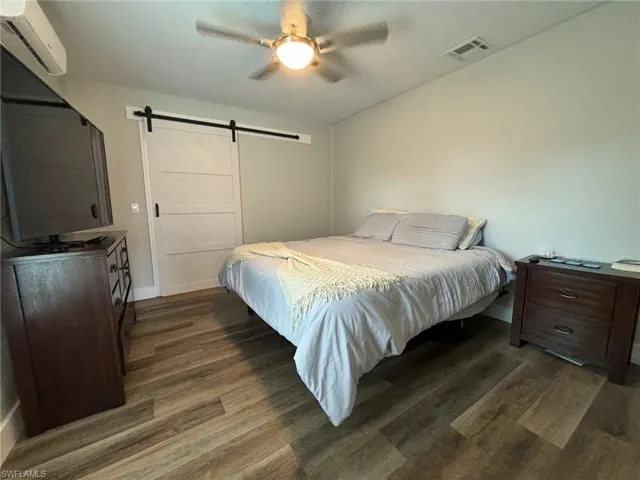 Bedroom with a wall mounted air conditioner, a barn door, dark wood-type flooring, and ceiling fan