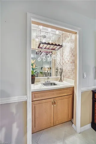 Recessed wet bar featuring light wood cabinetry, a stainless steel sink, tiled backsplash, and an overhead wine glass rack