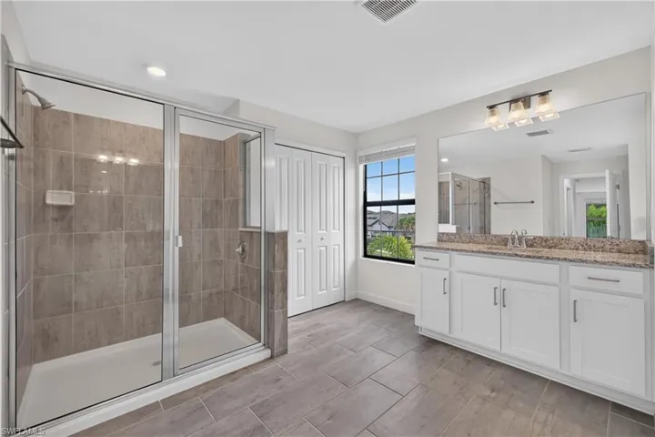 Spacious bathroom featuring a glass-enclosed shower with tile surround, a dual-sink vanity with stone countertop, tile flooring, and a window with blinds