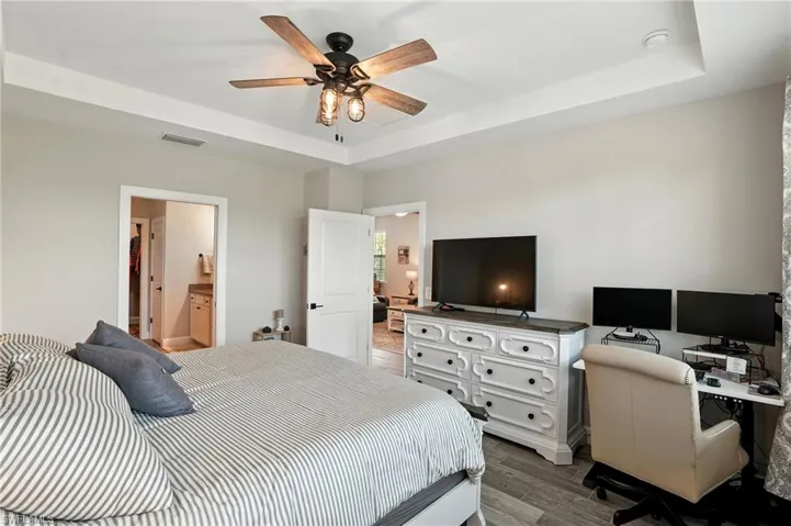 Bedroom with a raised ceiling, light wood finished floors, an office area, ensuite bath, and a ceiling fan