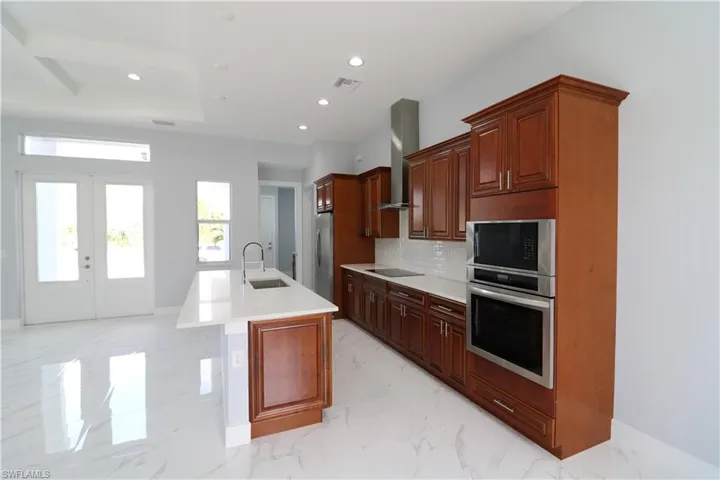 Kitchen featuring a center island with sink, stainless steel refrigerator, black electric stovetop, decorative backsplash, and wall chimney range hood