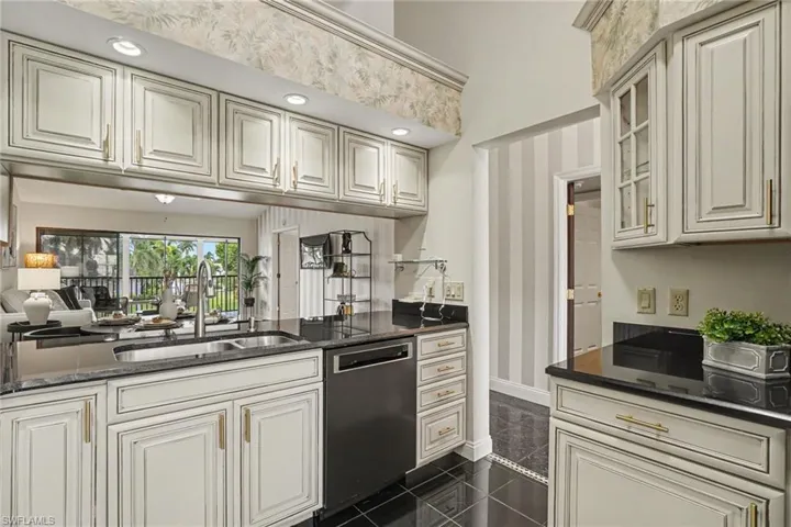 White cabinetry with gold-tone hardware, dark countertops, black tile flooring, and integrated stainless steel dishwasher