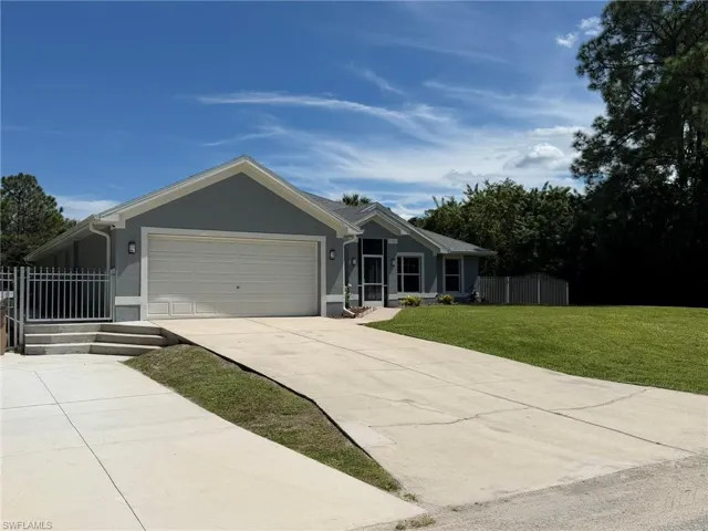 Gray stucco exterior with white trim, attached two-car garage, concrete driveway, front lawn, and mature trees
