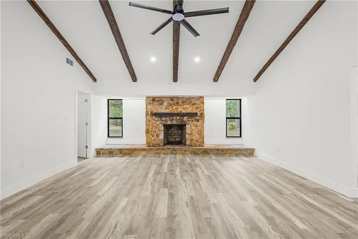 Spacious living area featuring a stone fireplace with a raised hearth, vaulted ceiling with exposed wood beams, and a black ceiling fan