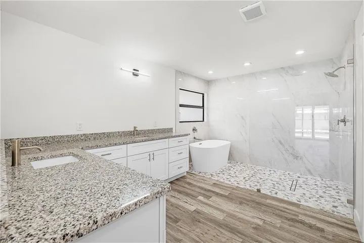 Spacious bathroom featuring a dual vanity with granite countertops, wood-finish flooring, a freestanding soaking tub, and a glass-enclosed shower with pebble tile flooring and large format tile walls