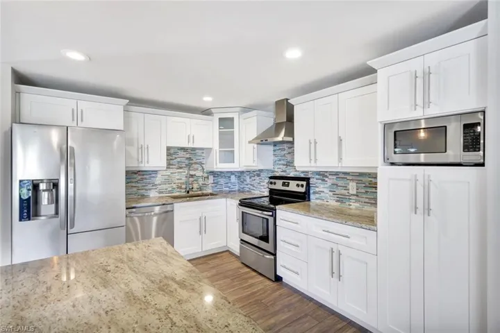 Kitchen featuring stainless steel appliances, light stone counters, wall chimney exhaust hood, white cabinets, and glass insert cabinets