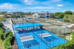 Recreational complex featuring multiple pickleball courts with blue surfacing, fenced tennis courts, shaded seating structures, and landscaped grounds