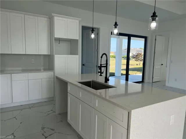 Kitchen featuring light stone counters, a kitchen island with sink, sink, decorative light fixtures, and white cabinetry