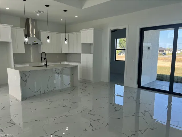 Kitchen featuring light stone counters, wall chimney exhaust hood, a kitchen island with sink, sink, and white cabinetry