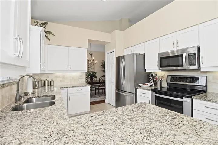 Kitchen with decorative backsplash, stainless steel appliances, light stone counters, white cabinets, and vaulted ceiling
