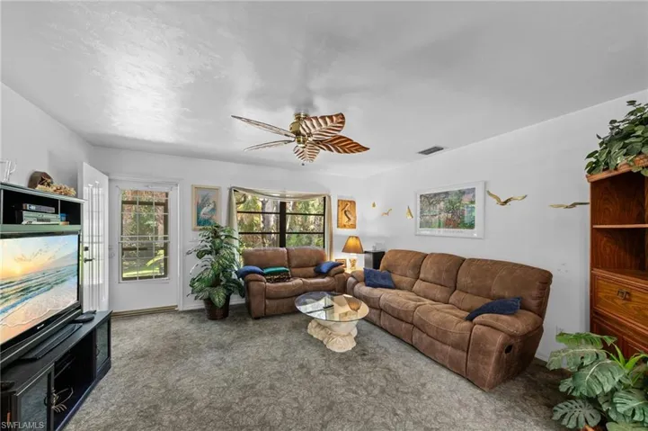 Living area featuring light gray wall-to-wall carpeting, white painted walls, and a ceiling fan with tropical-inspired blades
