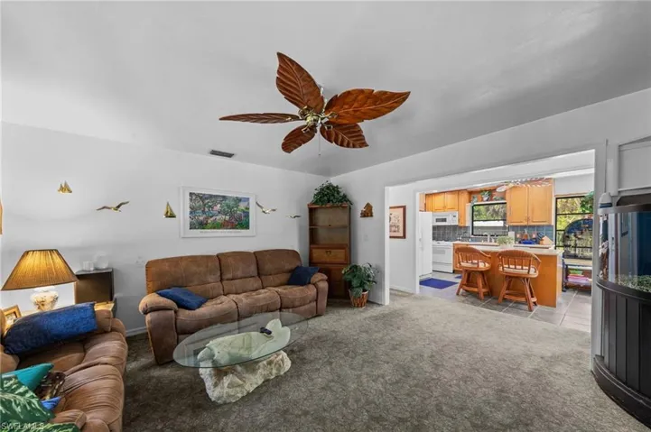 Open-concept living area with neutral carpeting, a ceiling fan featuring leaf-shaped blades, and an expansive doorway to the kitchen